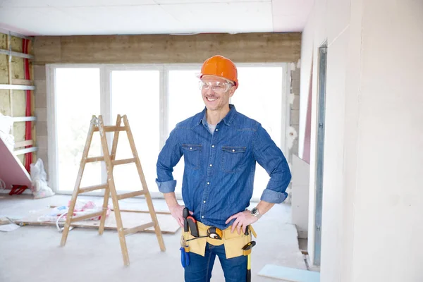 Portrait of thinking handyman wearing safety helmet and protective eye goggles while standing with hands on his hip at the construction site.