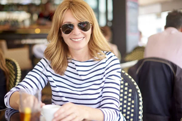Yakın çekim portre orta gülümseyen bir kafede oturan ve kahve içme kadın yaşlı. Güneş gözlüğü ve rahat kıyafetler giymiş ve rahatlatıcı çekici Olgun kadın.