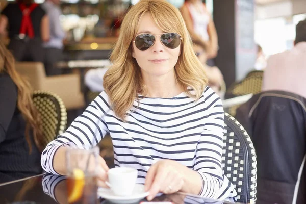 Orta gülümseyen portresi kafede oturan ve kahve içme kadın yaşlı. Güneş gözlüğü ve rahat kıyafetler giymiş ve rahatlatıcı çekici Olgun kadın.