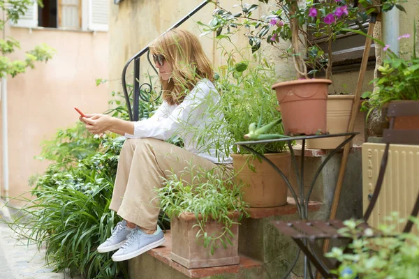Emin gülümseyen kadın Caddesi ve metin mesajlaşma yeşil bitkiler ile merdivende oturuyordu doz tam uzunlukta. 
