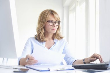Bazı evrak işlerini yapıyor ve ofis masa başında oturan ve çevrimiçi çalışıyorsanız dizüstü bilgisayarda yazarak orta yaşlı işkadını portresi.