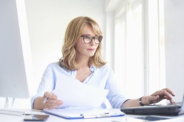 Bazı evrak işlerini yapıyor ve ofis masa başında oturan ve çevrimiçi çalışıyorsanız dizüstü bilgisayarda yazarak orta yaşlı işkadını portresi.