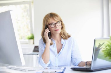 Dizüstü bilgisayarda çalışan office masasında otururken cep telefonu müvekkili ile konuşurken çekici olgun işkadını yakın çekim portresi.