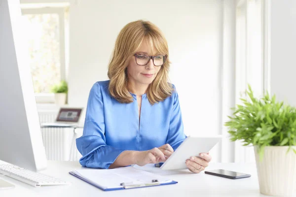 Portrait of mature executive businesswoman using digital tablet and doing some paperwork while sitting at her workstation.