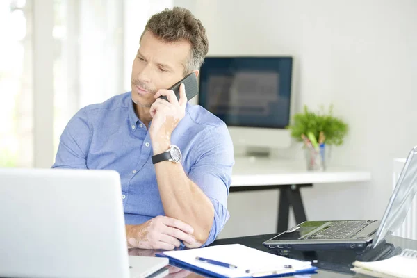 中年商人的肖像穿着衬衫 而坐在办公室和工作 专业的人使用他的手机 并与某人交谈 而在笔记本电脑工作 — 图库照片