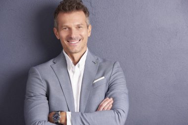Gri duvara ayakta ve kameraya bakarak katlanmış silah ile kendine güvenen bir orta yaşlı işadamı yakın çekim portre vurdu.