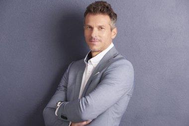 Gri duvara ayakta ve kameraya bakarak katlanmış silah ile kendine güvenen bir orta yaşlı işadamı yakın çekim portre vurdu.