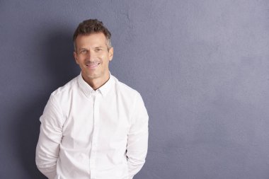 Gömlek giymiş ve kamera duran gri duvara bakarak yakışıklı orta yaşlı adam.