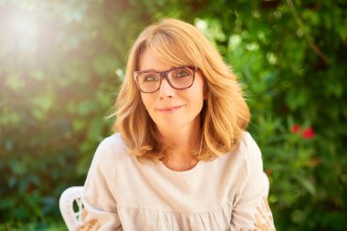 Rahat kıyafetler giymiş ve bahçede oturan ve rahatlatıcı kameraya bakarak mutlu orta yaşlı kadın görüntüsünü kırpılmış.