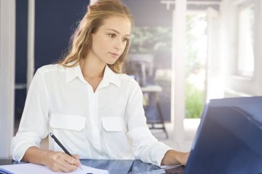 Güzel genç işkadını süre laptop önünde oturan ve iş projede çalışan kağıt üzerinde notlar yapma. 