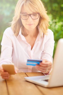 Orta yaşlı kadın cep telefonu ve kredi kartı kullanarak laptop önünde oturan ve online alışveriş. 