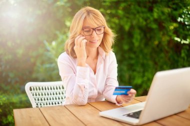 Orta yaşlı kadın onun banka kartı ve cep telefonu kullanarak laptop önünde oturan ve online bankacılık kadeh. 