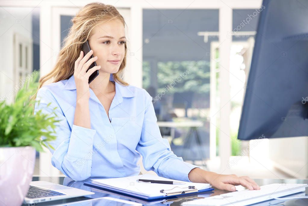Portrait of beautiful young assistant businesswoman making call while sitting at office desk and working on computer.
