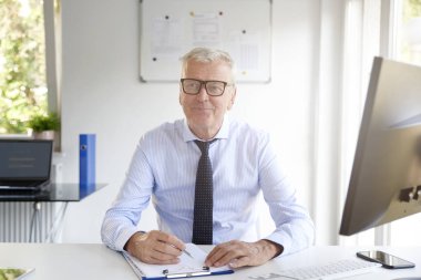 Ofis masa başında oturan ve bir kaç evrak kameraya bakıyor ve gülümseyen yaşlı işadamı portresi. 
