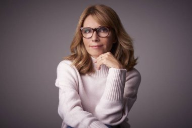 Yakın çekim studio kameraya bakarak ve açık gri renkli otururken gülümseyen orta yaşlı kadın portresi.