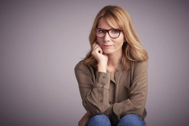 Studio portrait of confident middle aged woman sitting in front of light grey background, while looking at camera and smiling.  clipart