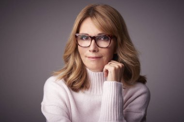 Yakın çekim studio kameraya bakarak ve açık gri renkli otururken gülümseyen orta yaşlı kadın portresi.