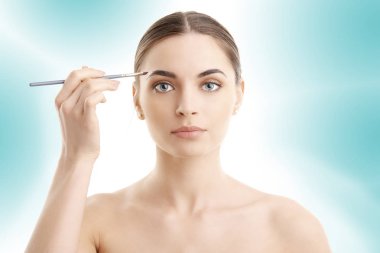 Güzel bir genç kadının onun kaş duran izole açık mavi renkli, makyaj uygulamak Studio vurdu. 