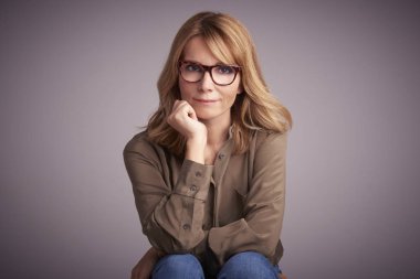 Kameraya bakarak ve gülümseyen açık gri renkli önünde oturan kendine güvenen orta yaşlı kadın portresi stüdyo. 