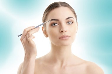 Güzel bir genç kadının onun kaş duran izole açık mavi renkli, makyaj uygulamak Studio vurdu. 