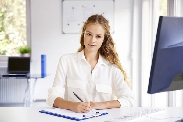 Piękna Młoda Kobieta Robiąc Notatki Siedząc Przy Biurku Przed Komputerem — Zdjęcie stockowe