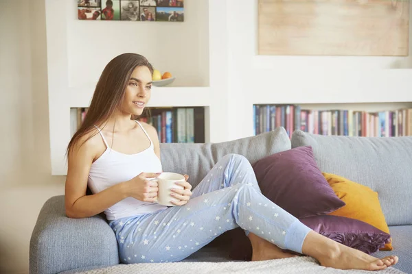 魅力的な若い女性のパジャマを着て 彼女のアパートでソファーでリラックスしながら彼女の朝のお茶を飲んでの全身ショット — ストック写真