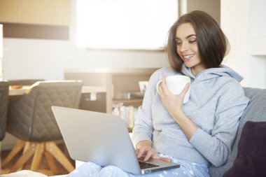 Atış onu laptop kullanarak ve evde kanepede otururken çay içme gülümseyen genç kadın.