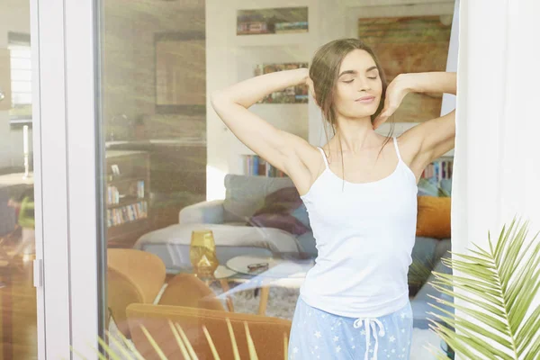 Pretty woman wearing pajamas and standing near window while stretching after waking up.