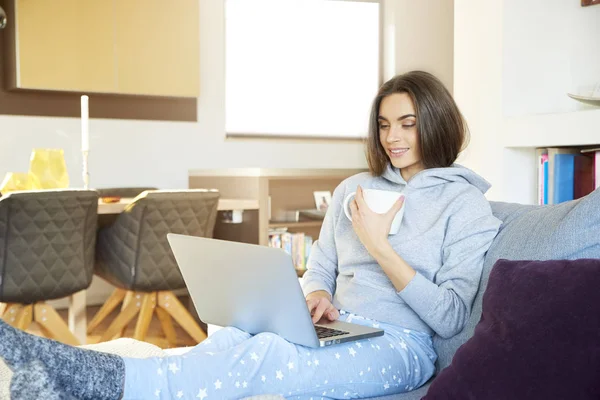 彼女のラップトップを使用して 自宅でソファに座ってお茶を飲んで笑顔の若い女性のショット — ストック写真
