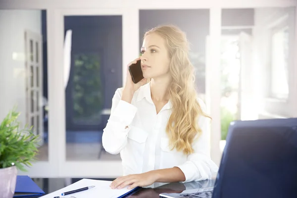 美しい若い実業家のオフィスの机に座っていると彼女の携帯電話で誰かと話しての肖像画 — ストック写真