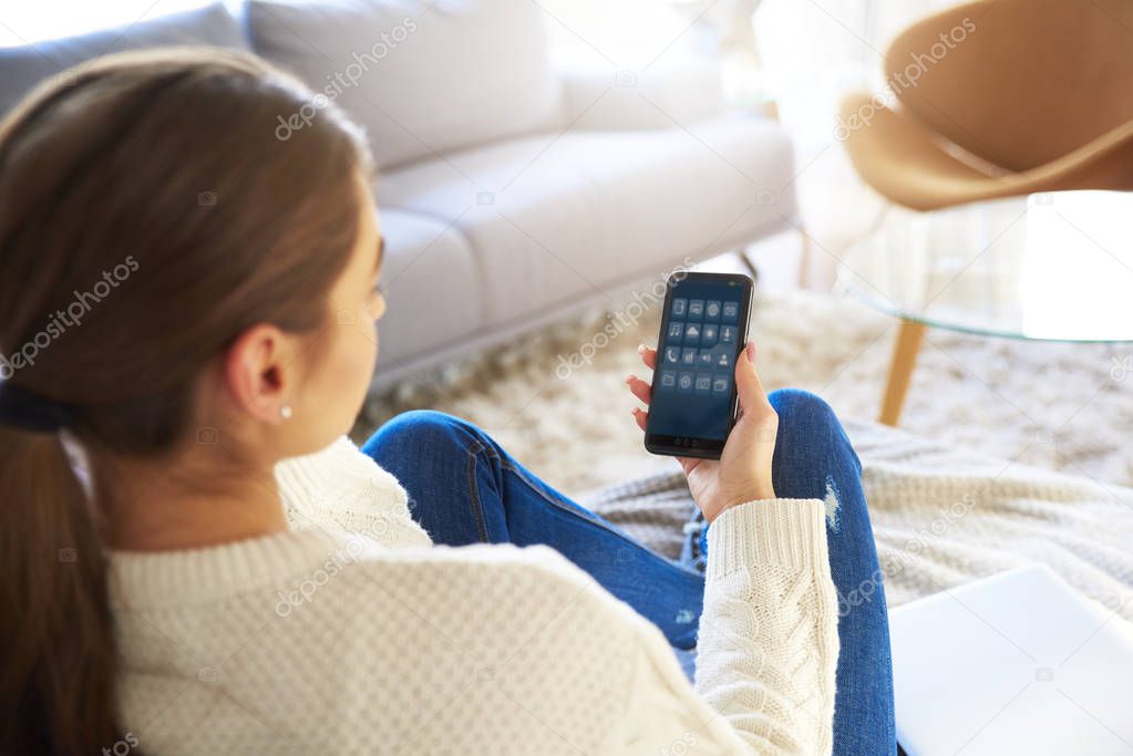 Rear view shot of woman holding mobile phone in her hand and using it while relaxing on sofa at home. 