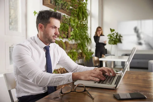 自信的销售人员穿着衬衫和领带 而坐在笔记本电脑前 在办公桌打字的肖像 站在幕后的无法辨认的女商人 — 图库照片