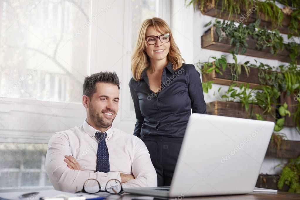 Shot of young financial businessman sitting at office desk while executive businesswoman standing next to him and working on laptop. Teamwork in the office. 
