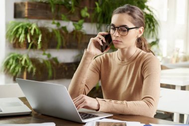Onu laptop ofis masası, arkasında oturan ve biriyle ofiste çalışırken cep telefonu üzerinde konuşurken güzel genç iş kadını portresi. 