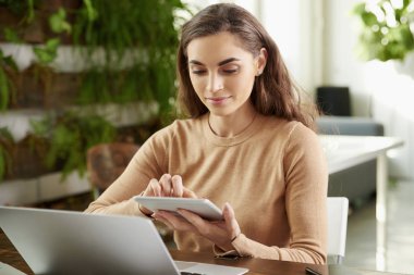 Ofis masa başında oturan ve çevrimiçi çalışıyorsanız genç işkadını holding dijital tablet onun elinde gülümseyen portresi.