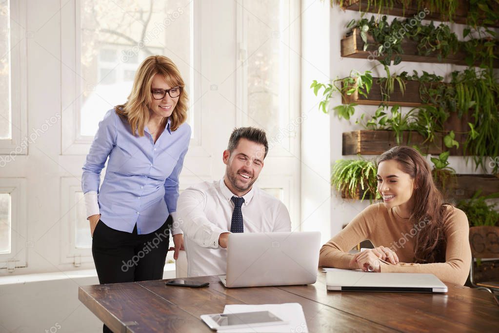 Group of business people working together on business project.  Young secretary woman and financial advisor businessman sitting in front of laptop while middle aged executive professional woman standing behind them and giving advice.