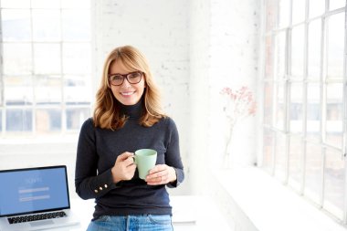 Portrait shot of attractive casual businesswoman holding mug in her hand while standing at office desk and relaxing.  clipart