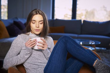 Kanepede oturan ve rahatlatıcı kupa onun elinde tutan güzel genç kadın portre vurdu.