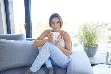 Tam uzunlukta atış pijama giymiş ve koltukta evde dinlenirken kupa onun elinde tutan genç kadın. 