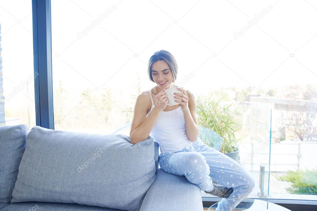 Full length shot of young woman wearing pajamas and holding mug in her hand while relaxing on sofa at home. 