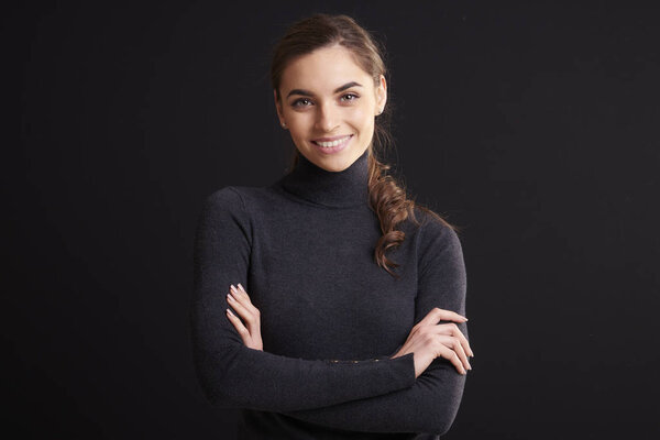 Крупным планом студийный портрет красивой молодой женщины в свитере с рулонной шеей и смотрящей в камеру, стоя со скрещенными руками на темном фоне
. 