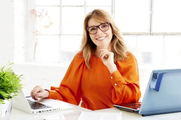 陽気な成熟した実業家彼女のノート パソコンの背後にあるオフィスの机に座っていると 新しいプロジェクトに取り組んでの肖像画のショット — ストック写真