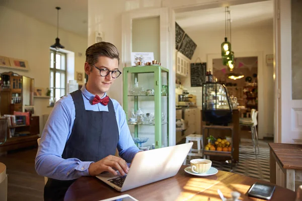 年轻人穿着围裙 坐在办公桌前 在咖啡店里处理笔记本电脑的照片 小型企业 — 图库照片