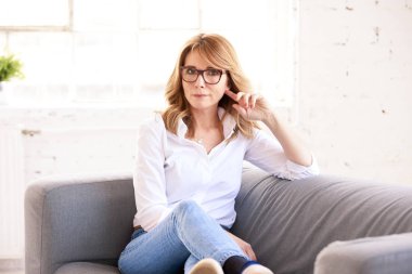 Kameraya bakarken ve gülümserken kanepede dinlenen kendine güvenen orta yaşlı kadının tam uzunlukta çekilmiş fotoğrafı. 