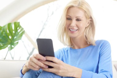 Onun cep telefonu ve metin mesajlaşma koltukta evde dinlenirken kullanan gülümseyen bir portre sarışın kadın. 