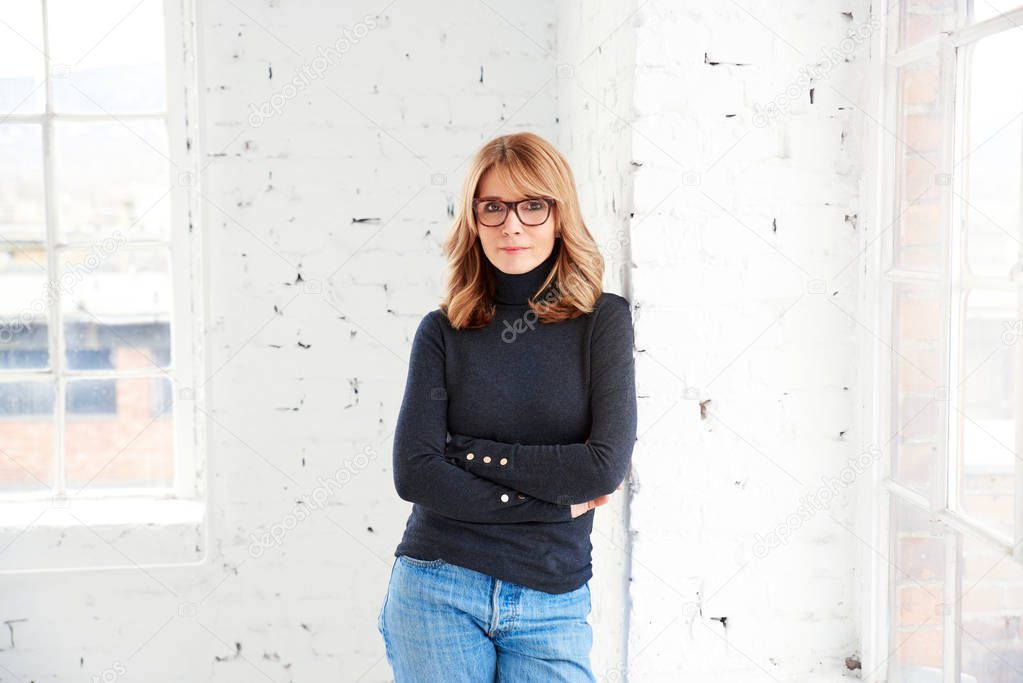 Portait of attractive middle aged woman wearing roll neck sweater and jeans while relaxing at the window and looking at camera.