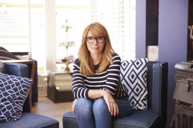 Evde kanepede otururken ve rahatlatıcı rahat giysiler giyen gülümseyen orta yaşlı kadının portre çekimi. 