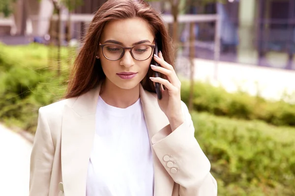 オフィスビルの間の通りを歩いている間 彼女の携帯電話で誰かと話している若いビジネスウーマンの肖像画 — ストック写真
