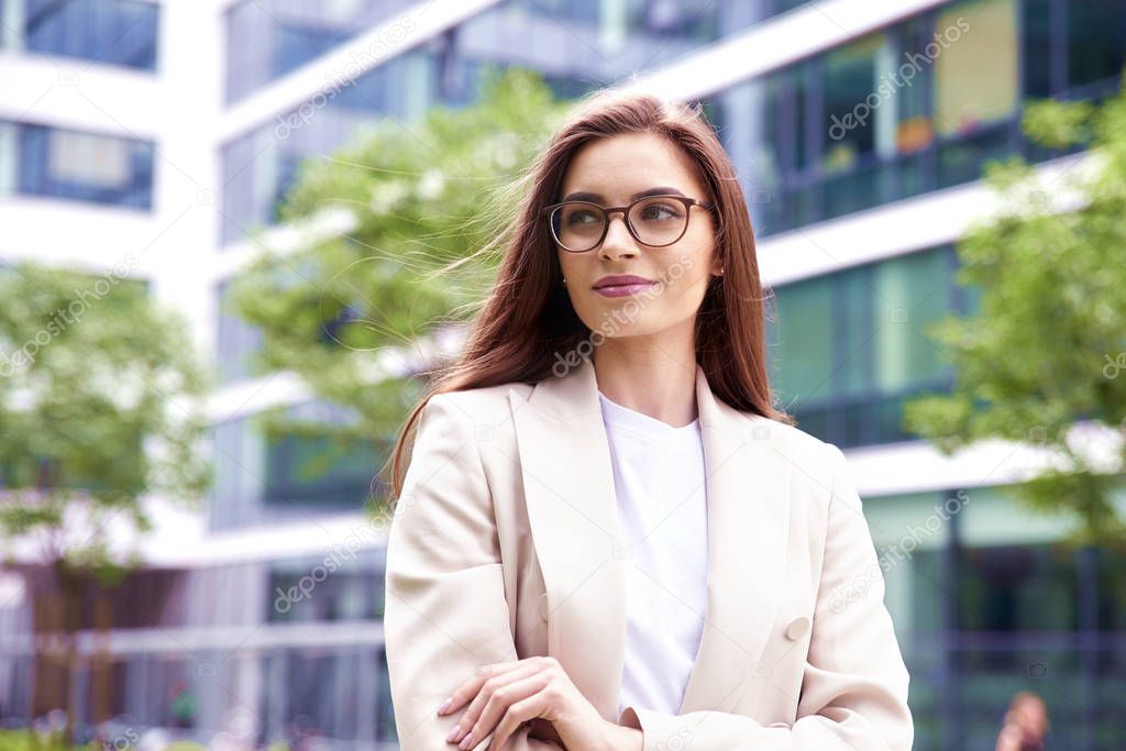 Shot of beautiful young businesswoman wearing blazer and eyeglasses while standing on the street with folded arms. 