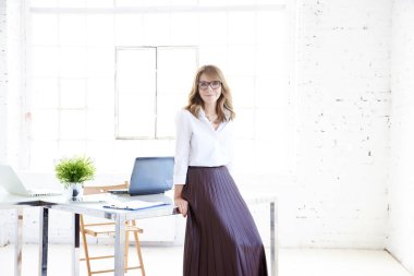 Çekici orta yaşlı iş kadını tam uzunlukta çekim kamera bakarak ve ofis masasında dururken gülümseyerek. 